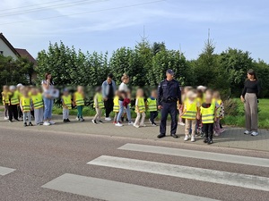 przed oznakowanym przejściem dla pieszych stoi policjant z grupą dzieci i ich opiekunkami. Dzieci mają na sobie kamizelki odblaskowe