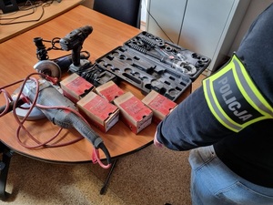 na stole leżą: szlifierka, wkrętarka, skrzynka z narzędziami, kartonowe opakowania. Obok stoi policjant służby kryminalnej