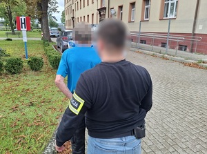 na tyłach budynku komendy Policji stoją policjant kryminalny, który przytrzymuje stojącego przed nim zatrzymanego mężczyznę
