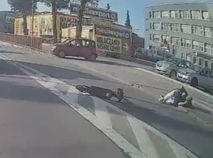 na jezdni leży mężczyzna. Kilka metrów dalej na jezdni leży hulajnoga. Na drodze dla rowerów przodem do jezdni, w odległości kilkunastu metrów od leżącego na jezdni mężczyzny, stoi czerwony osobowy samochód
