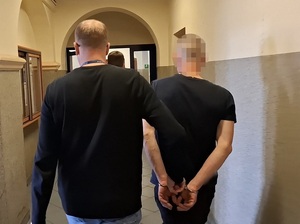nieumundurowany policjant prowadzi korytarzem mężczyznę, który ma ręce skute za plecami kajdankami
