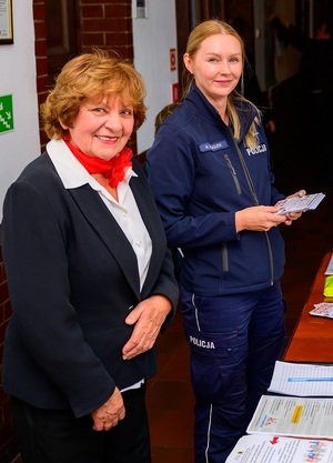 w sali przy stole z ulotkami stoją: uśmiechnięta seniorka i policjantka