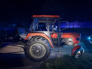 pora nocna. Na drodze stoi stary, zniszczony czerwony ciągnik rolniczy Ursus. Nie ma szyb ani fragmentu drzwi. Przed nim stoi policyjny radiowóz