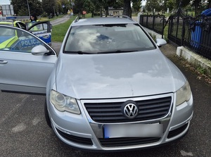 przy bramie posesji stoi srebrny VW Passat z otwartymi drzwiami od strony pasażera. Za nim stoi oznakowany policyjny radiowóz, a przy drzwiach radiowozu policjantka