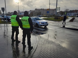 na chodniku przed przejściem dla pieszych stoją dwaj policjanci. Przez pasy przechodzi kobieta. Przed pasami znajduje się samochód osobowy. Pogoda deszczowa, pochmurno