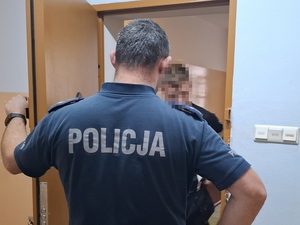 w celi stoi zatrzymany mężczyzna. Za nim stoi policjant służby dyżurnej, który przytrzymuje otwarte drzwi celi