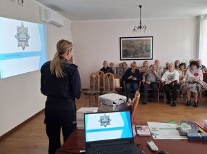 w sali naprzeciw siedzącym w rzędach seniorów stoi policjantka. Po jej lewej stronie na ścianie wyświetla się prezentacja