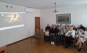w sali w rzędach siedzą na krzesłach seniorzy. Po ich prawej stronie na ścianie wyświetla się film profilaktyczny