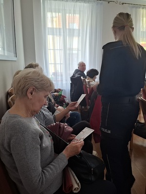 policjantka rozdaje siedzącym w rzędach seniorom ulotki profilaktyczne