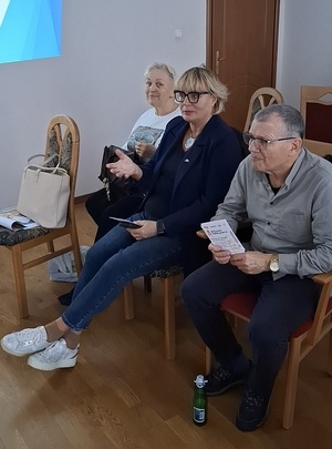 w sali na krzesłach siedzą seniorzy. Jedna z seniorek przemawia