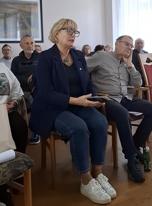 w sali na krzesłach w rzędach siedzą seniorzy. Jedna z seniorek przemawia