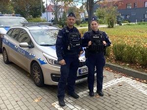 przed oznakowanym radiowozem stoi dwoje policjantów w umundurowaniu ćwiczebnym: policjant w stopniu sierżanta i policjantka w stopniu starszego posterunkowego